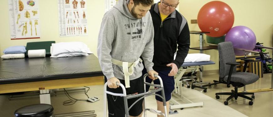 A student stands beside a man using a walker in a physical therapy setting